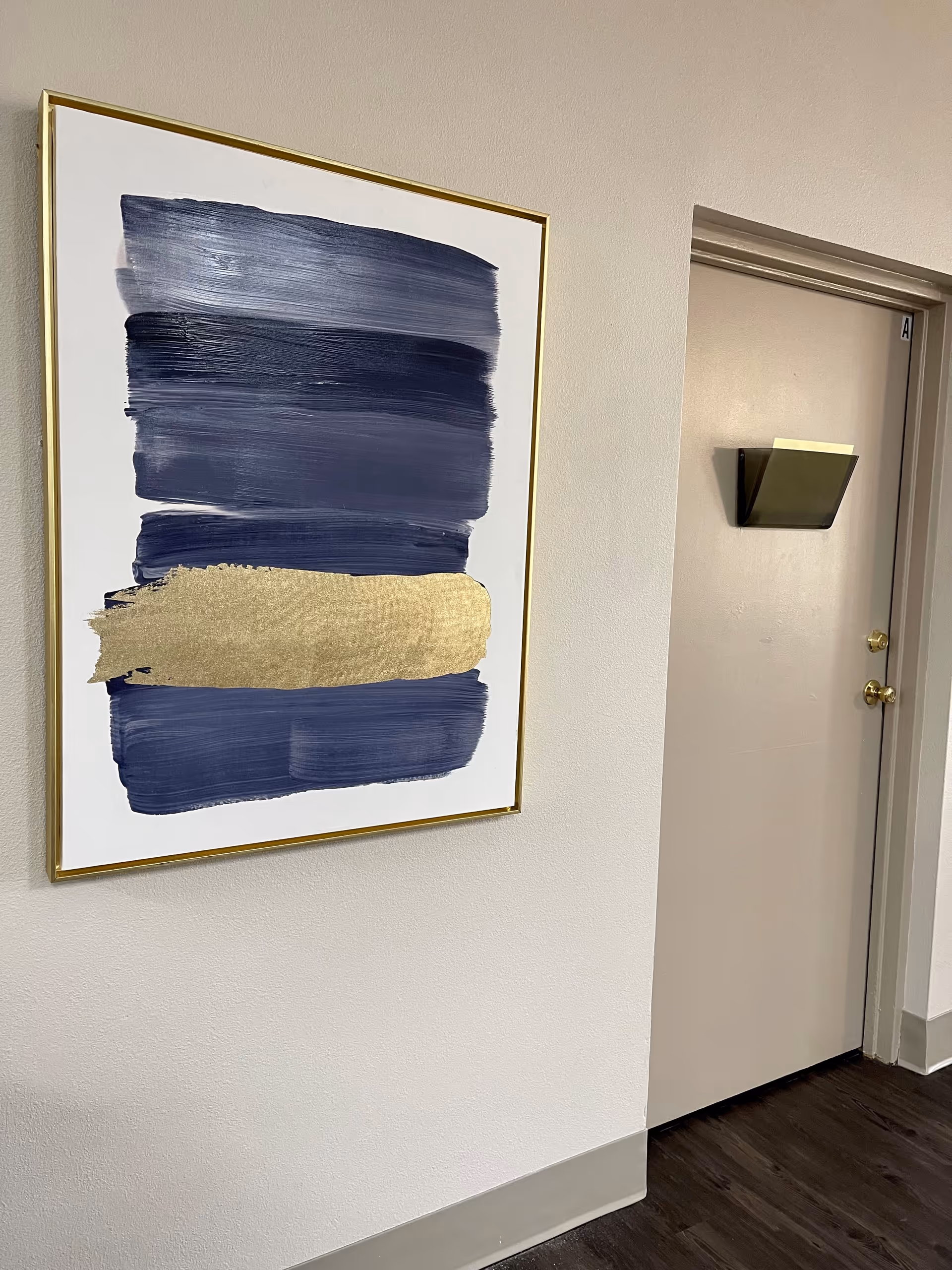 A hallway wall with a framed abstract painting featuring broad horizontal brush strokes in dark blue and gold. Next to the painting is a closed beige door with a small black wall-mounted holder and two gold-colored door knobs.