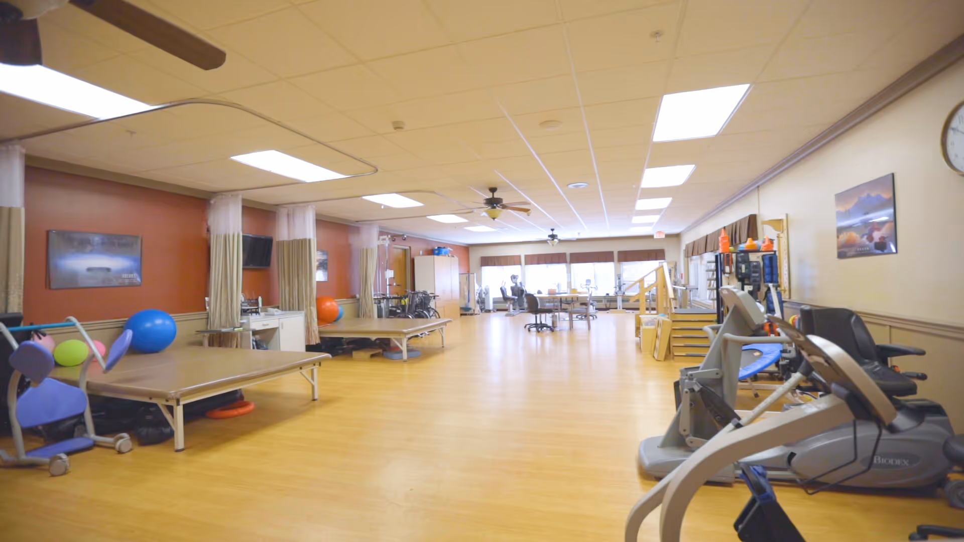 A spacious physical therapy or rehabilitation room with exercise equipment including stationary bikes, therapy tables, exercise balls, and parallel bars. The room has a wooden floor, ceiling fans, large windows letting in natural light, and a clock on the wall.