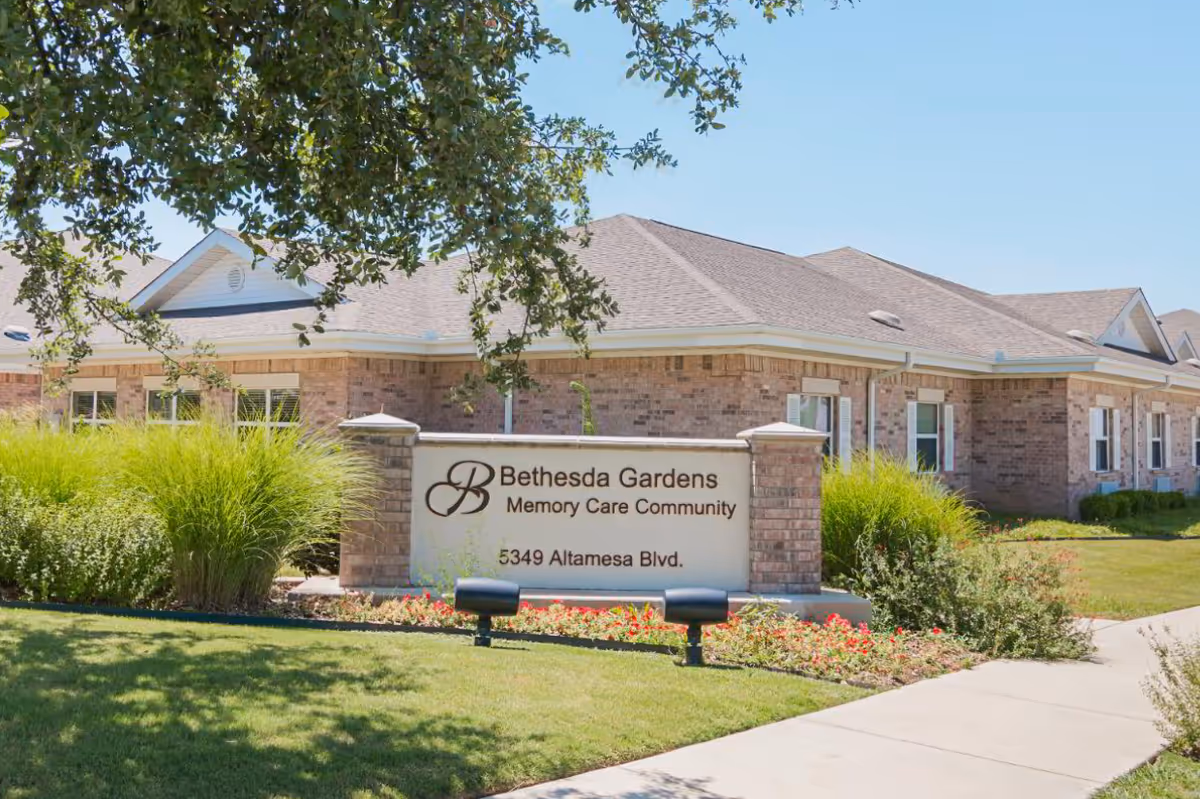 Exterior view of Bethesda Gardens Memory Care Community building with a brick facade, surrounded by green bushes and a well-maintained lawn. A large sign in front displays the facility name and address: 5349 Altamesa Blvd.