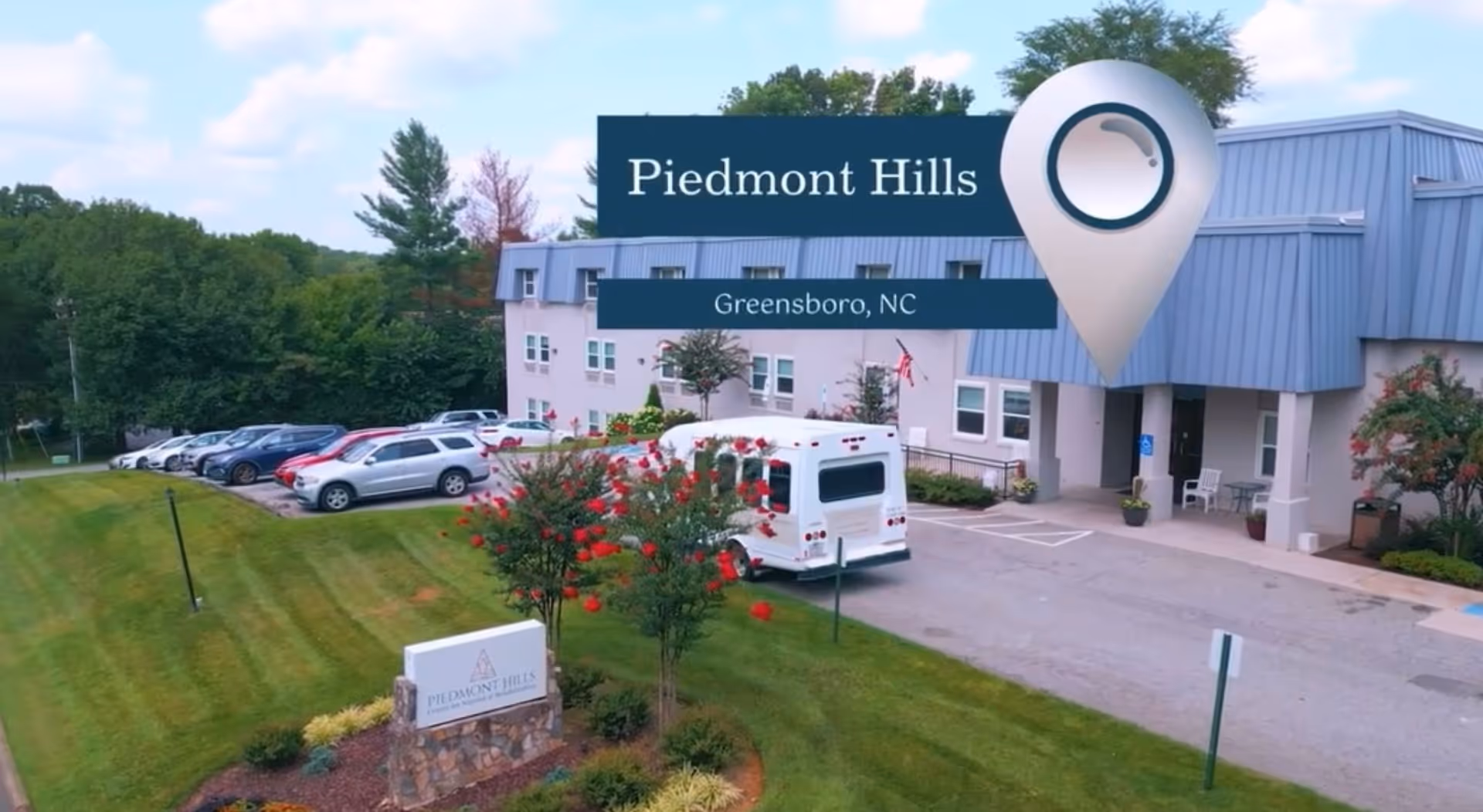 Exterior view of Piedmont Hills Center for Nursing and Rehabilitation in Greensboro, NC, showing the building entrance, a parking lot with several cars, a white shuttle bus, and landscaped greenery with trees and flowers.