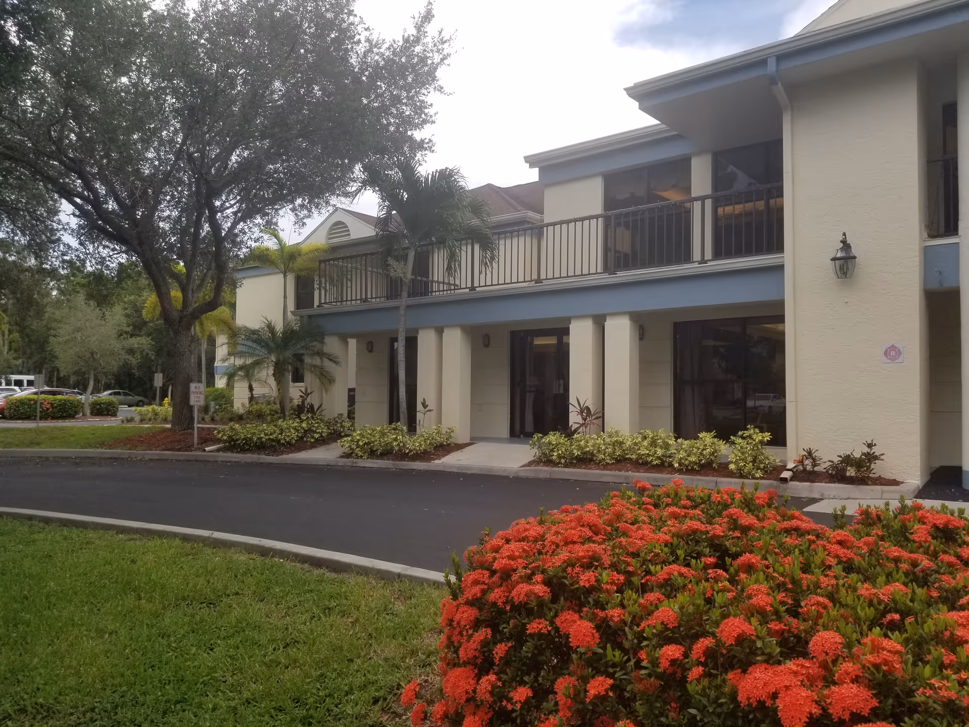 Exterior view of a two-story senior living facility building with beige walls and blue trim. The building has large windows and a balcony on the upper floor. There are palm trees and other greenery around the building, with a well-maintained garden featuring red flowers in the foreground. A paved driveway curves in front of the building.