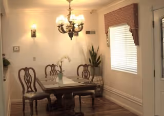 A traditional dining room with a wooden table and chairs beneath a chandelier, a window with blinds and valance, and a potted plant.