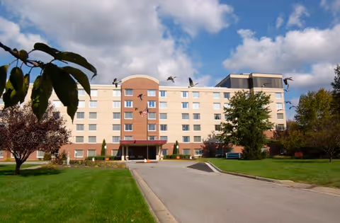 Front exterior of a multi-story senior living building with a driveway, green lawn, trees, and birds flying overhead.