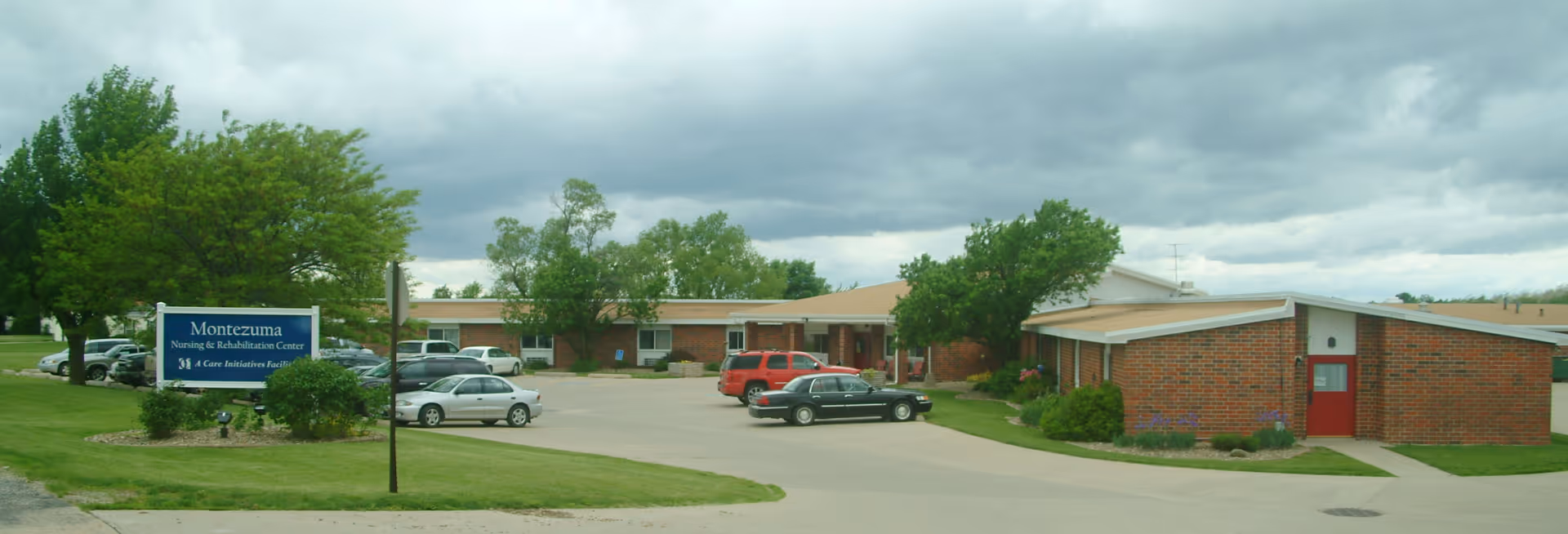 Front view of a single-story brick nursing and rehabilitation building with a parking lot, several cars, a lawn and a blue sign reading 'Montezuma' under a cloudy sky.
