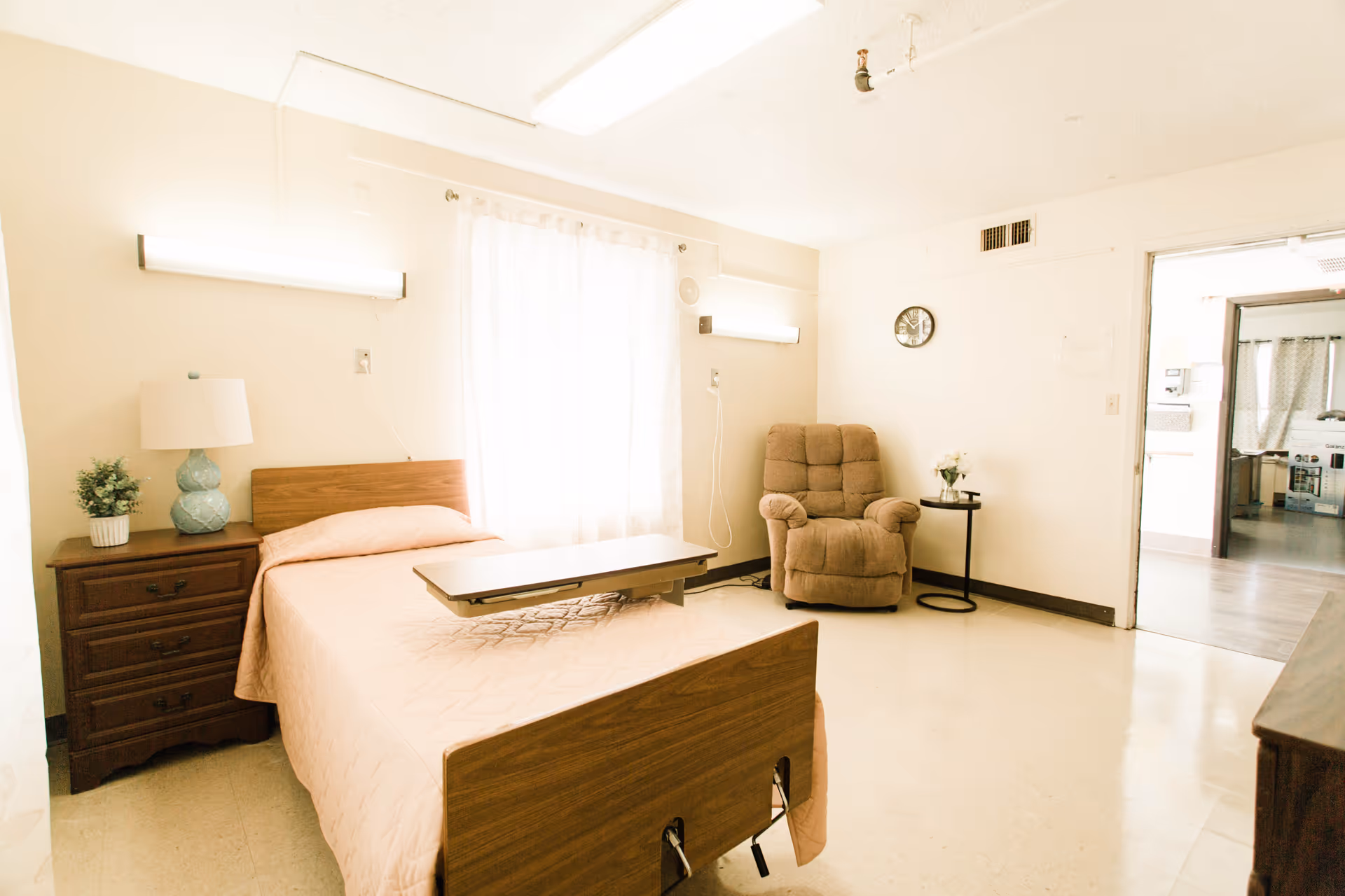 A simple furnished bedroom with bed, nightstand, lamp, recliner, and a small table.