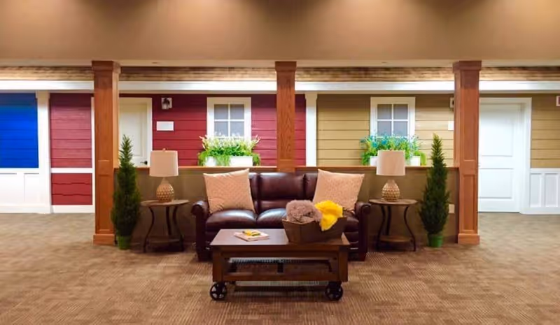 A cozy seating area with a dark brown leather sofa adorned with two light-colored pillows. In front of the sofa is a wooden coffee table with a basket containing folded blankets and a book. On either side of the sofa are small round tables with matching lamps and potted plants. Behind the seating area is a wall designed to look like the exterior of small cottages with colorful siding in blue, red, and beige, white doors, and windows with flower boxes. The floor is carpeted in a neutral tone.