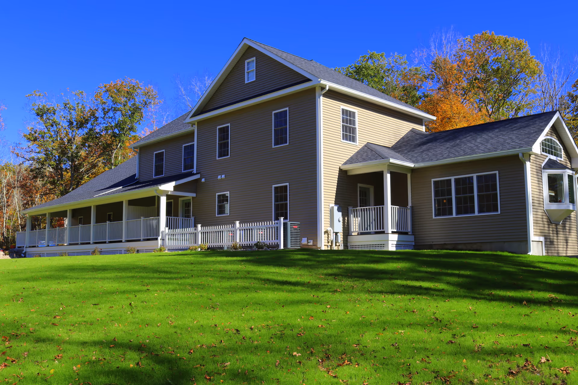 Light-brown two-story house with a wraparound porch on a green lawn under a clear blue sky.
