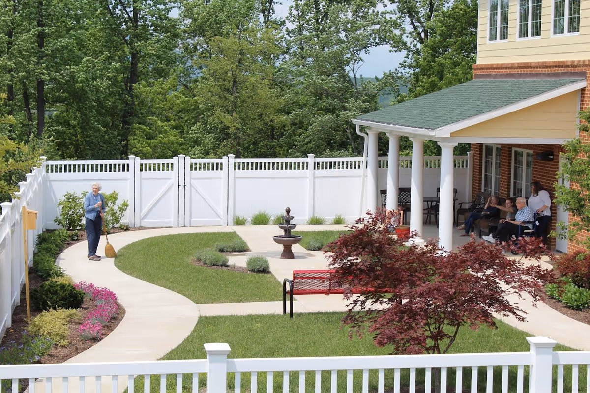 Outdoor garden area at Hidden Springs Senior Living with a paved walking path, green lawn, a small fountain, and various plants and flowers. An elderly woman is sweeping the path while a group of elderly people and a caregiver sit and chat under a covered patio attached to a brick building.