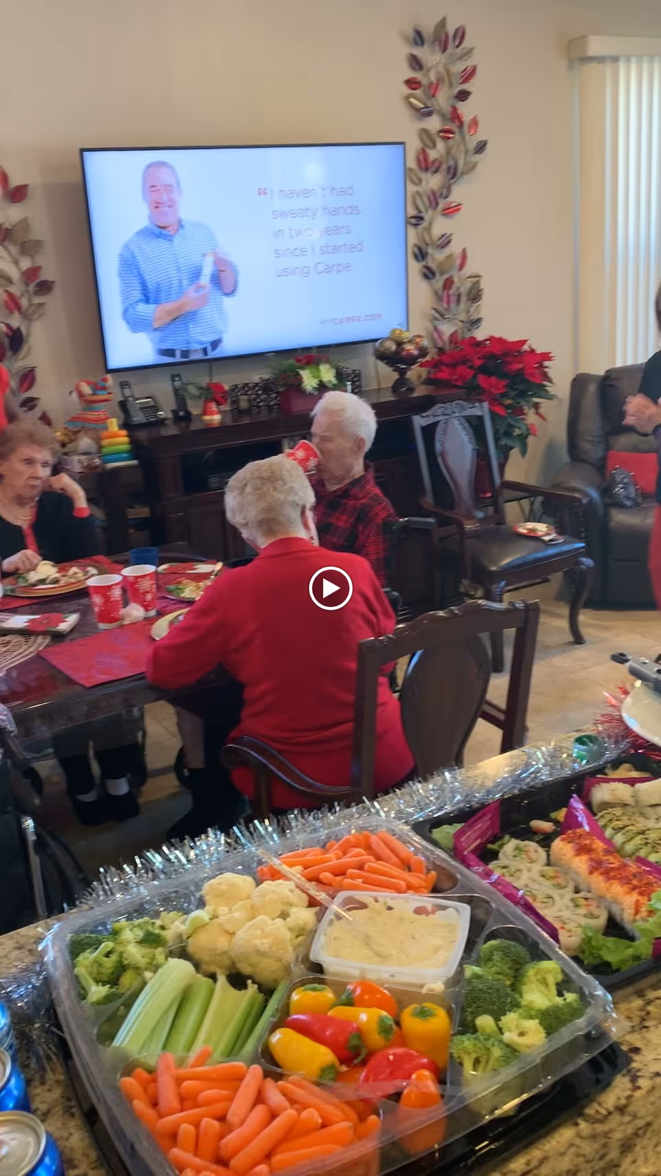 A group of elderly people sitting around a dining table enjoying a meal together in a decorated room. In the foreground, there is a tray of fresh vegetables including carrots, celery, cauliflower, broccoli, mini bell peppers, and a container of dip. A television screen on the wall displays an advertisement featuring a man and text about using Carpe for sweaty hands. The room is decorated with holiday-themed items including poinsettias and garlands.