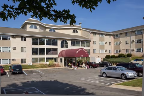 Exterior view of Promenade Senior Living building with maroon entrance canopy and parked cars.