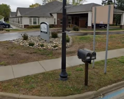 Exterior view of a senior living facility with sign, mailbox, and lamppost along sidewalk.