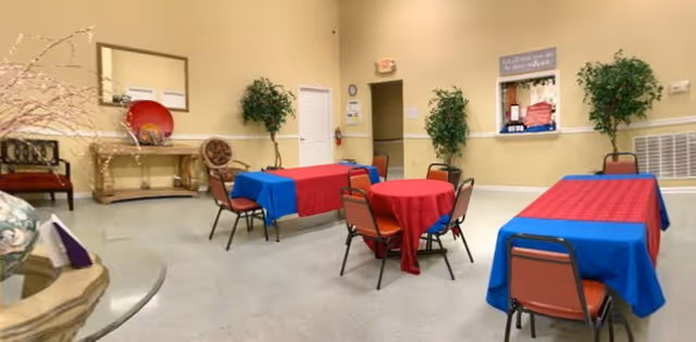 A common area in a senior living facility with several tables covered in red and blue tablecloths, surrounded by chairs. The room has beige walls, two artificial plants, a mirror above a console table, and a wall sign with an inspirational quote.