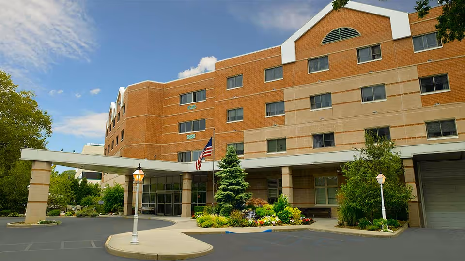 Front entrance of a multi-story brick senior living facility with a covered driveway, landscaping, and an American flag.