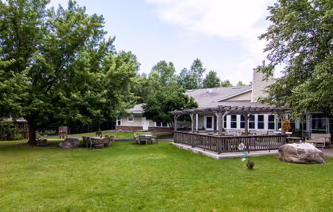 Outdoor area of a senior living facility with green grass, several trees, wooden benches and tables, large rocks, and a building with a covered patio and multiple windows under a partly cloudy sky.