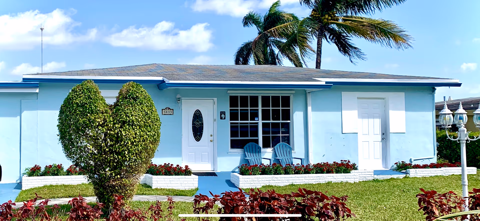 Light-blue, single-story facility exterior with a front porch, two chairs, and palm trees.