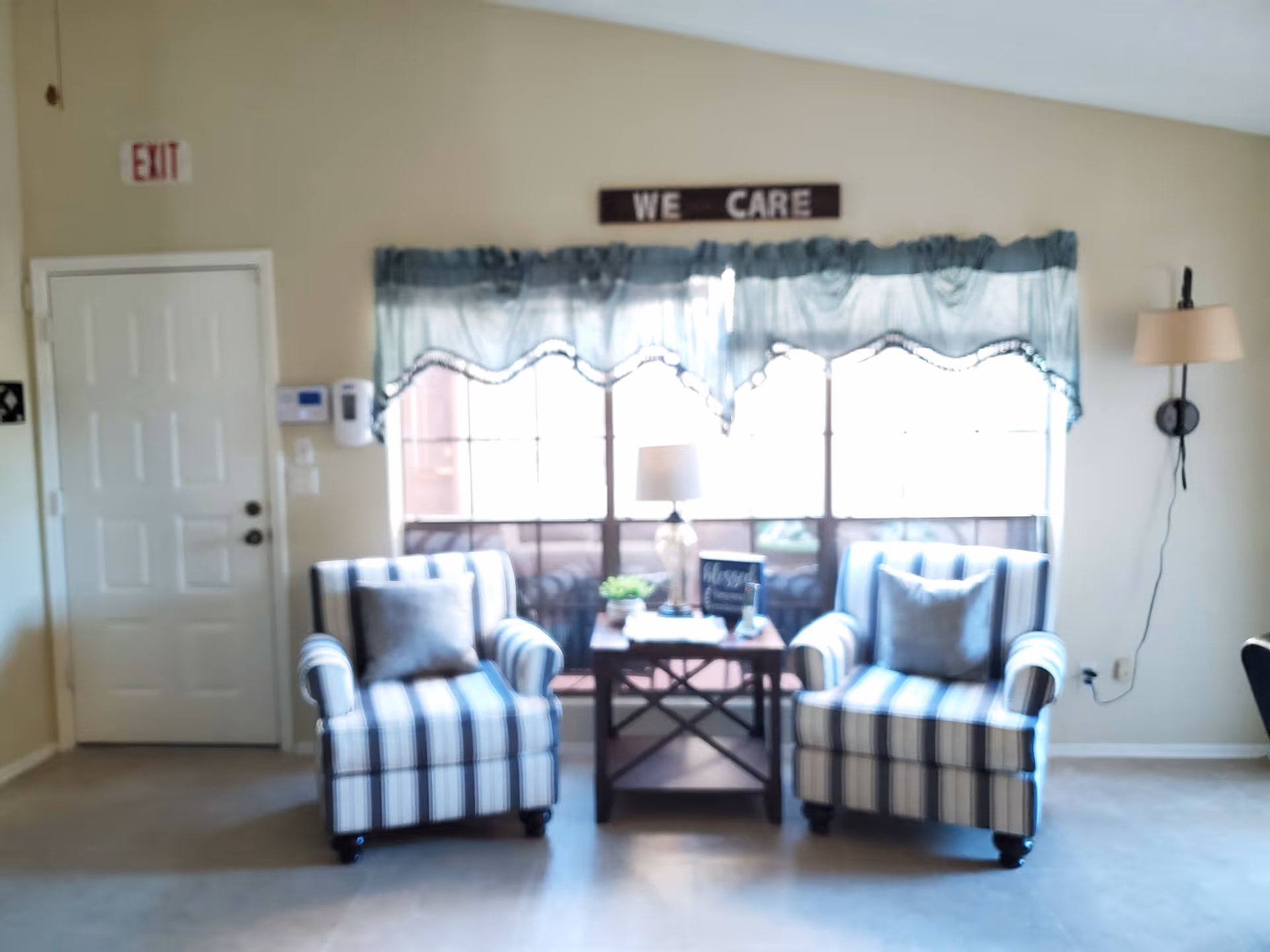 A cozy seating area with two striped armchairs facing forward, each with a gray pillow. Between the chairs is a small wooden table with a lamp, a small plant, and a decorative sign. Behind the seating area is a large window with blue curtains and a sign above that reads 'WE CARE'. To the left is a white door with an exit sign above it, and to the right is a wall-mounted lamp.