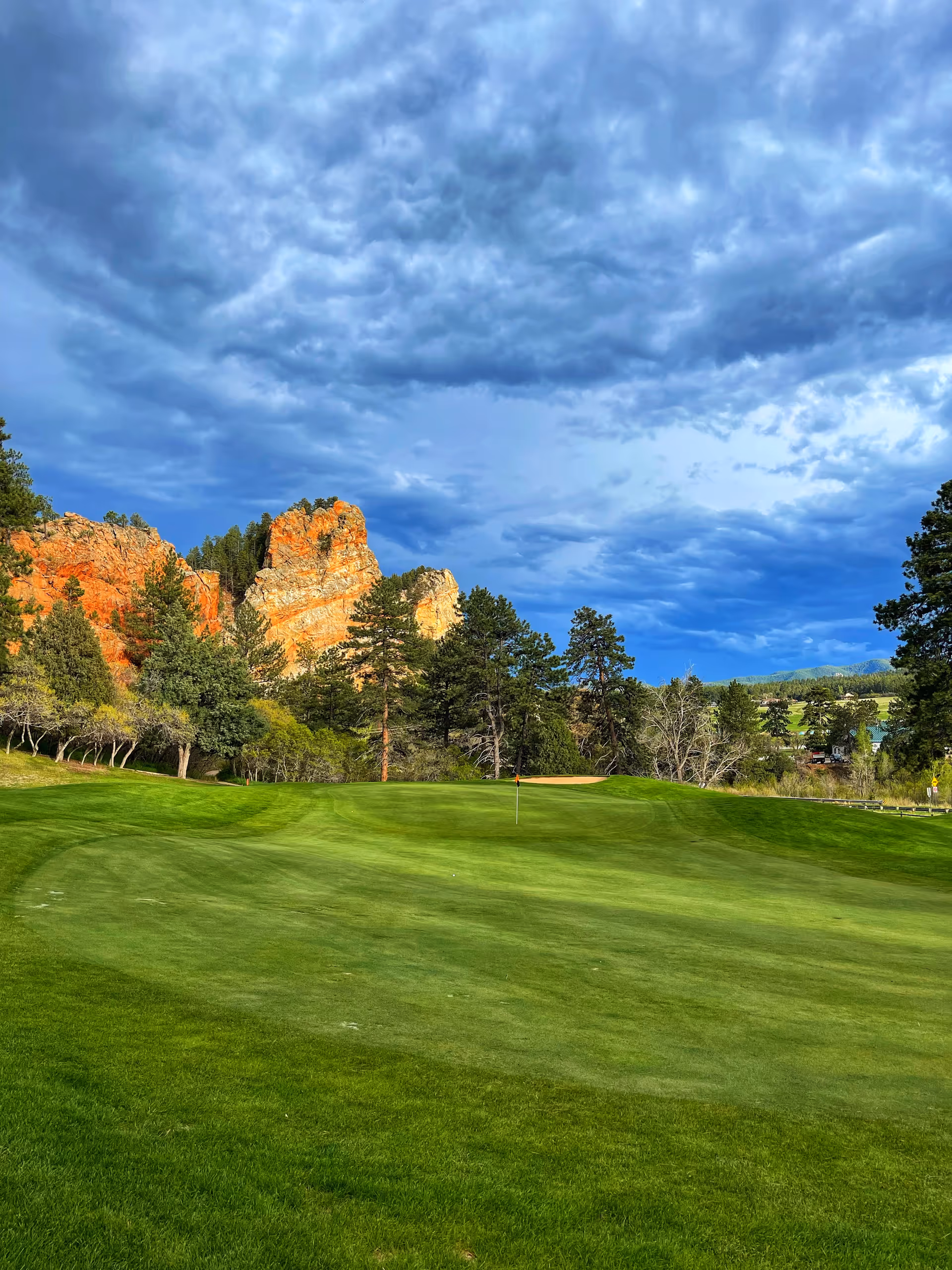 Lush green golf course fairway with trees, orange cliffs, and a cloudy blue sky.