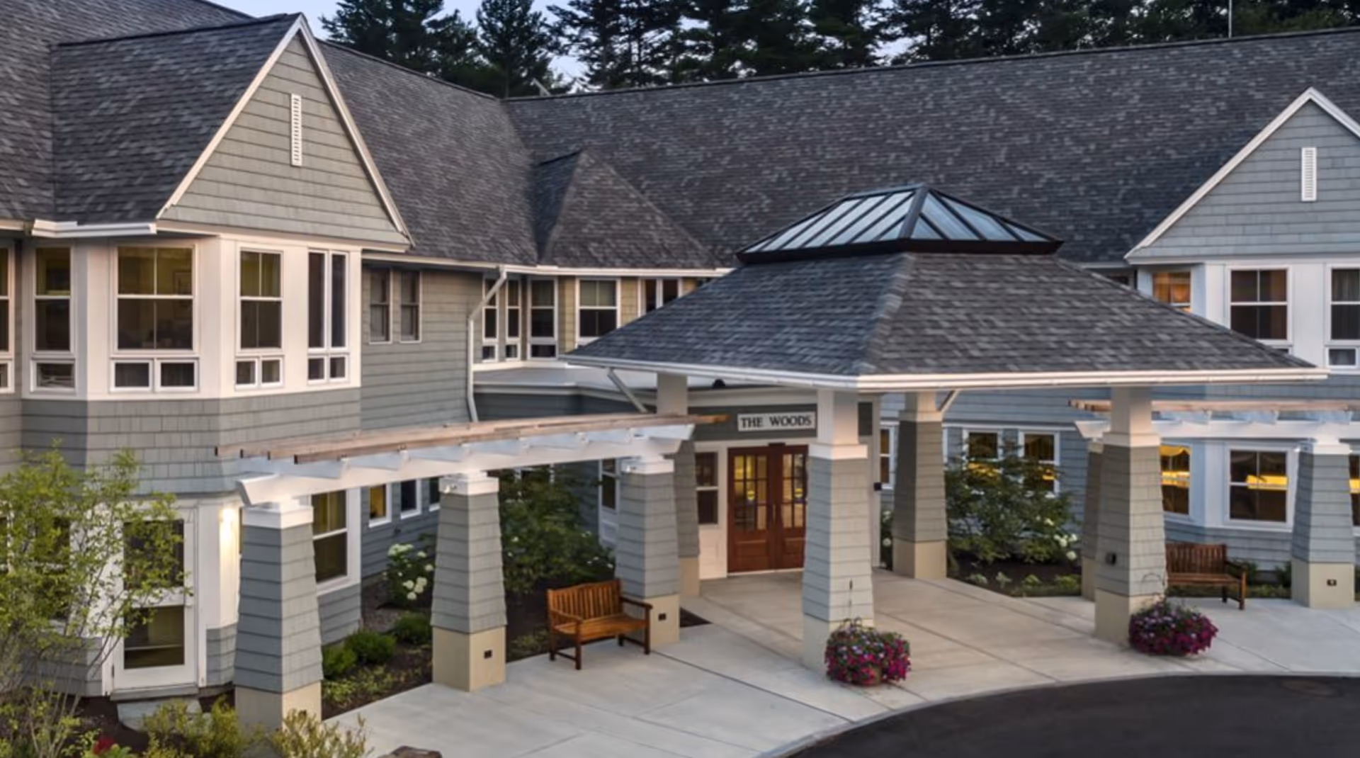 Exterior view of a senior living facility entrance with a covered drop-off area supported by columns, benches on either side, and landscaping with flowers and shrubs. The building has gray siding, white trim, and multiple windows under a shingled roof.