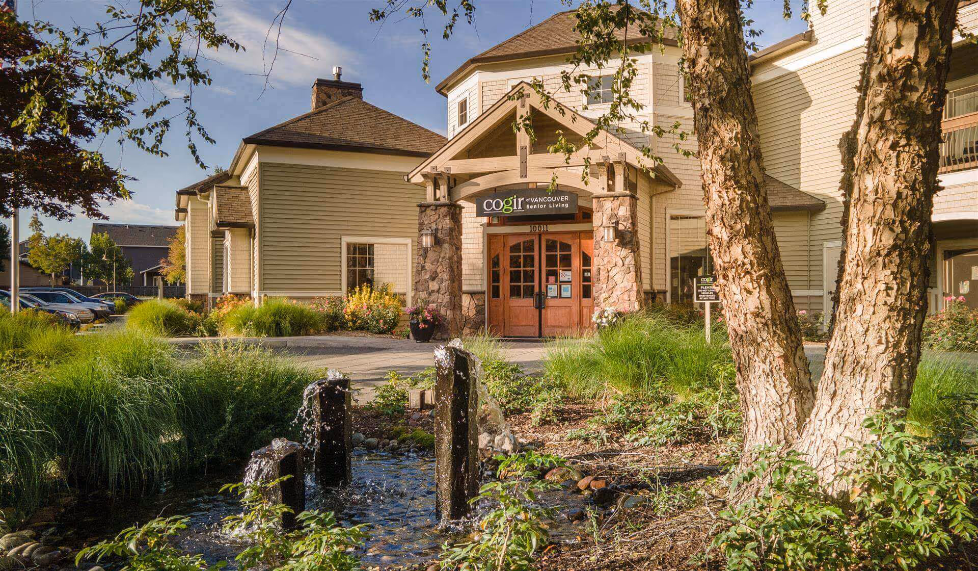 Front entrance of a senior living building with landscaped grounds and a small water feature.