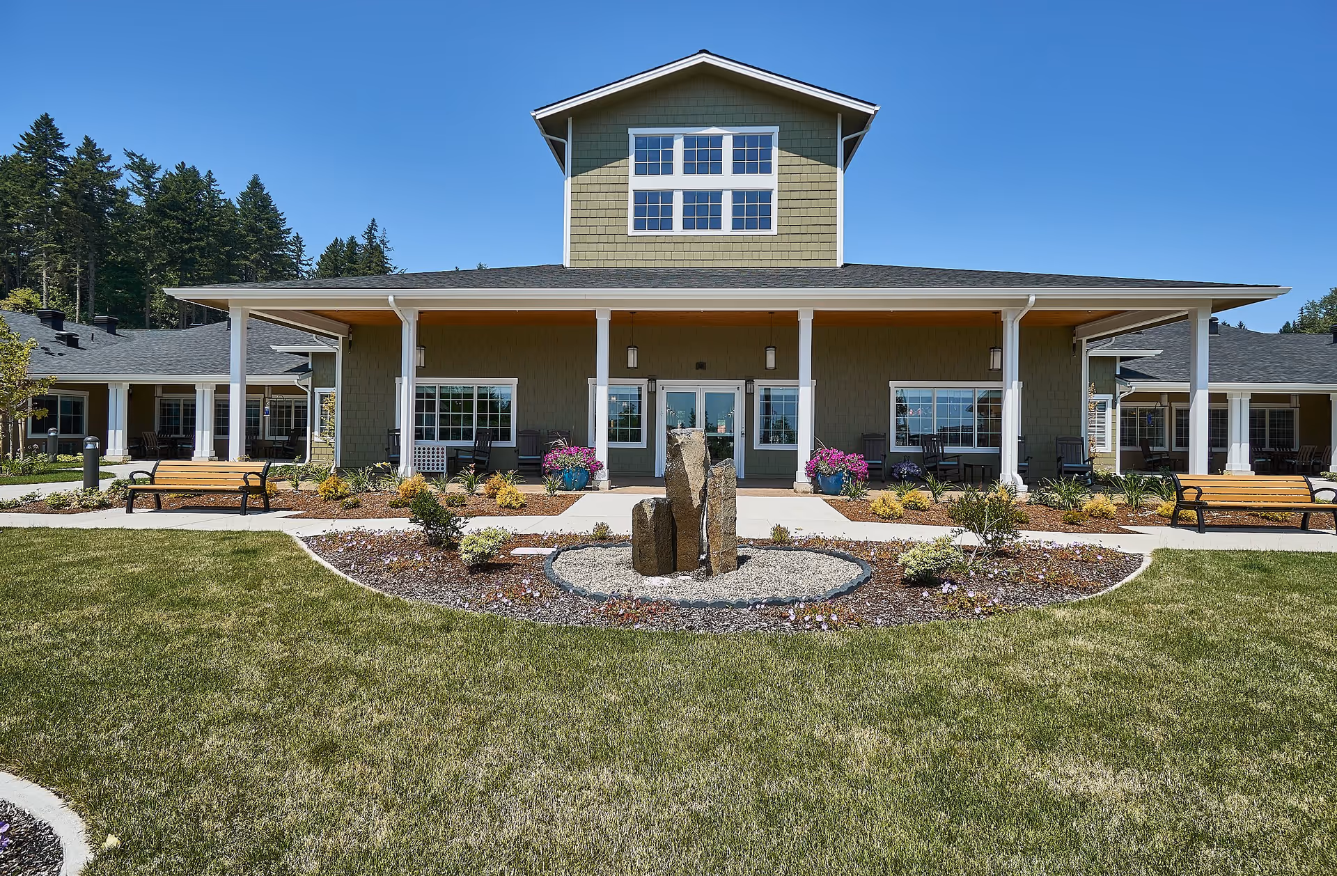 Front exterior of a senior living building with a covered porch, benches, landscaped lawn and a central stone fountain.