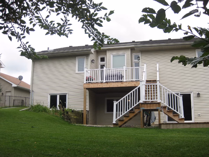Two-story beige siding house with a wooden deck and white railing in a grassy yard