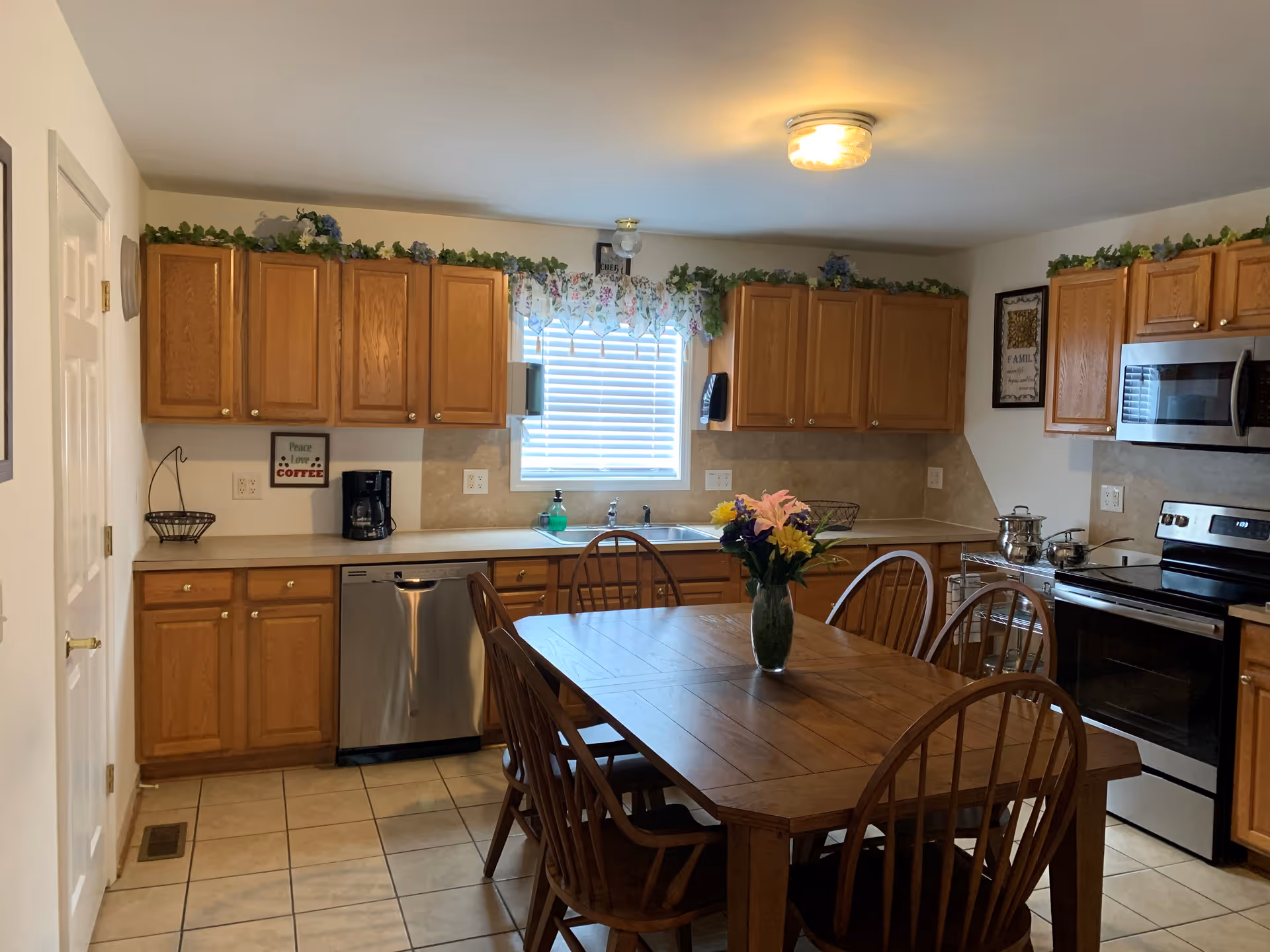 A kitchen with wooden cabinets, a stainless steel dishwasher, stove, and microwave. There is a wooden dining table with six chairs in the center, and a vase with colorful flowers on the table. A window with floral curtains and greenery decorates the top of the cabinets.