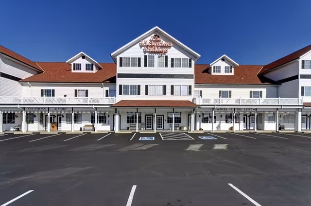 Large white senior living facility exterior with red roof and parking in front.
