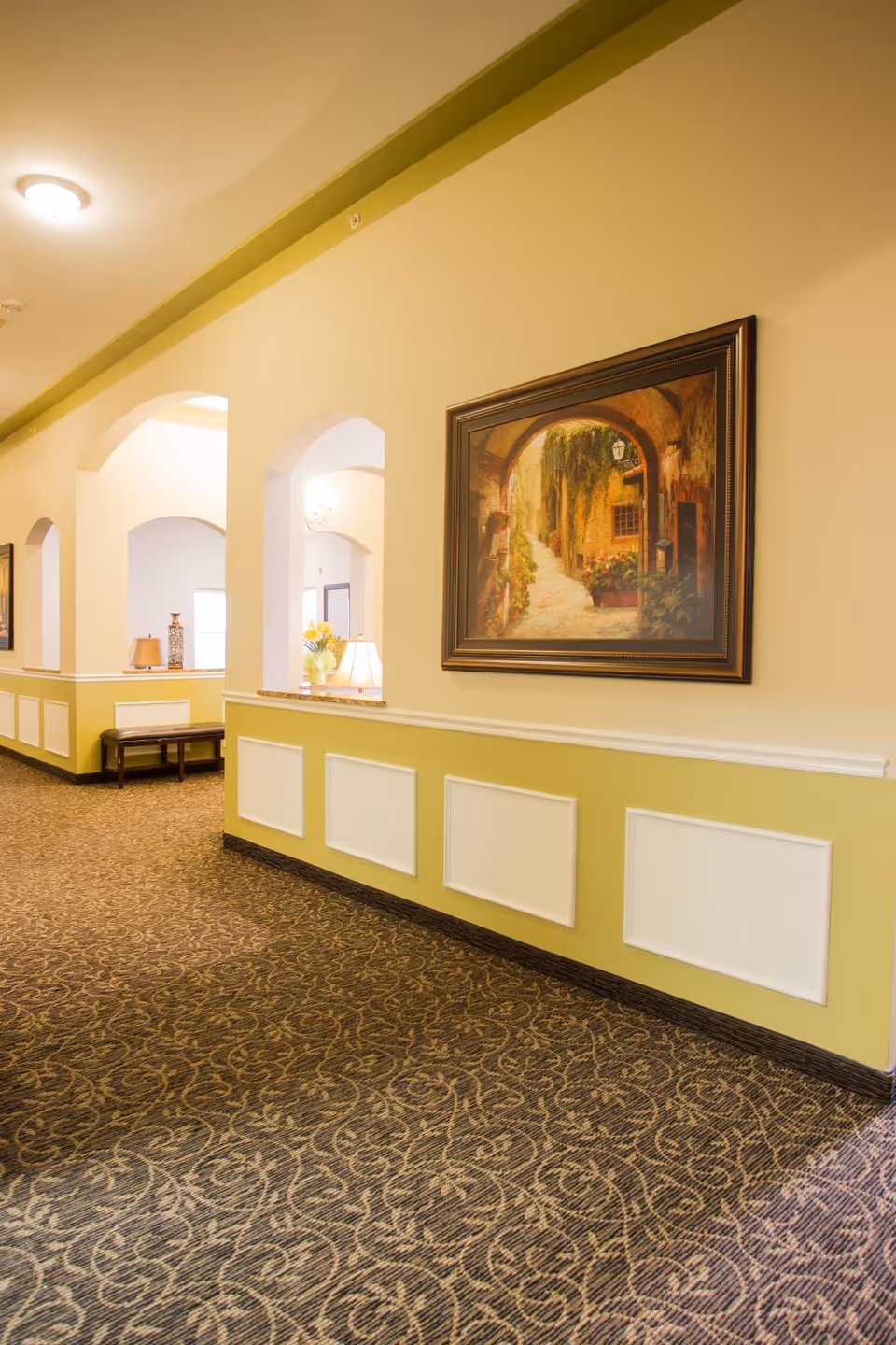 Interior corridor with arched openings, lamps, paintings, and patterned carpet.
