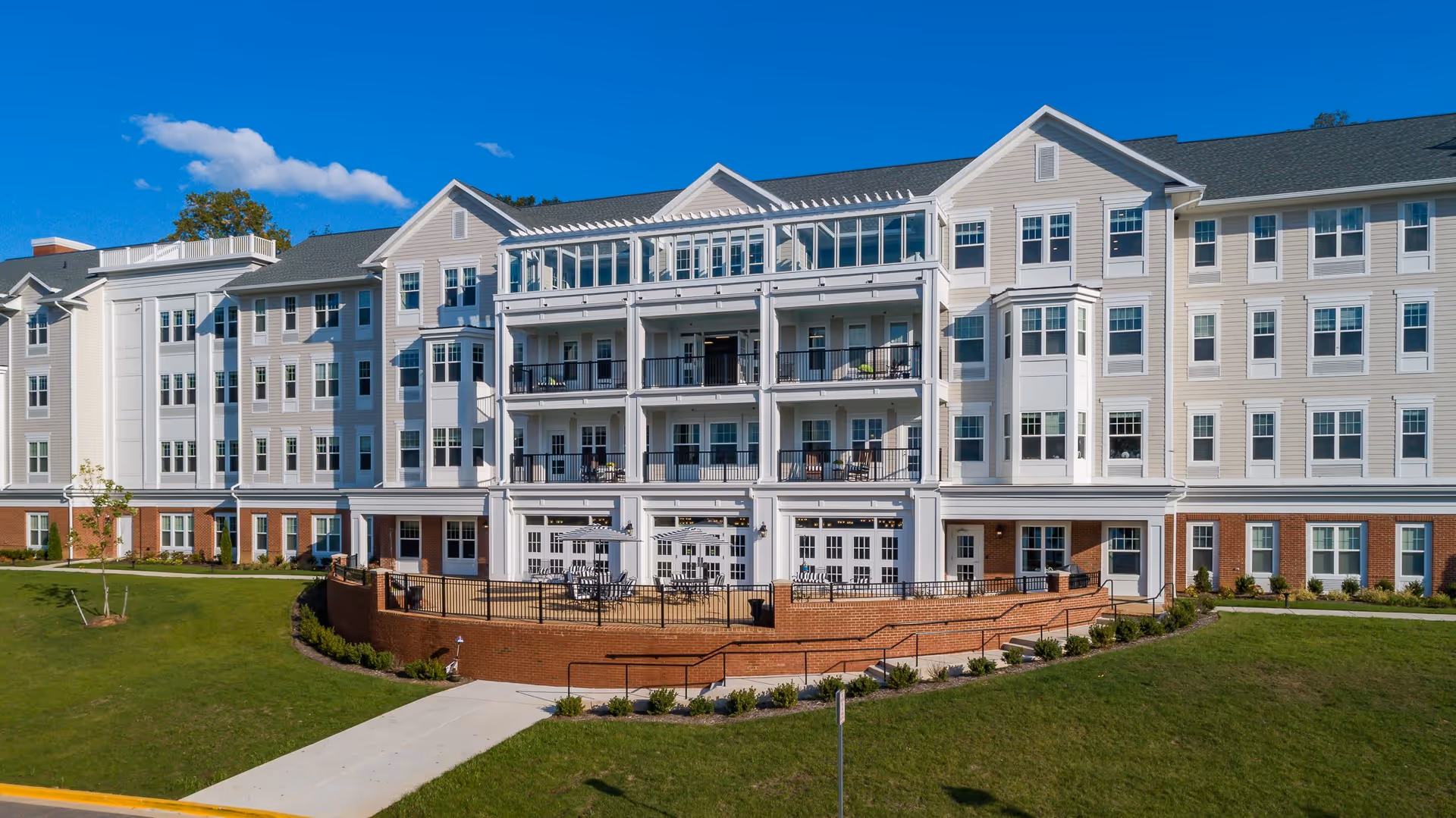 Exterior view of a large, multi-story senior living facility building with multiple balconies and a patio area with tables and umbrellas. The building has white siding with brick accents on the lower level and is surrounded by a well-maintained lawn and landscaping under a clear blue sky.