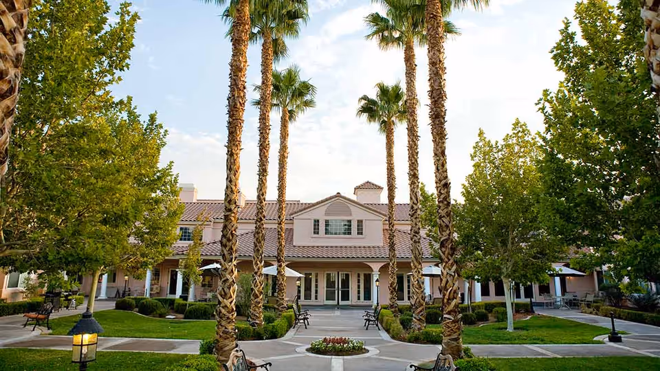 View of the exterior courtyard of a senior living facility with tall palm trees, green lawns, benches, and a building with a tiled roof and multiple windows in the background under a partly cloudy sky.