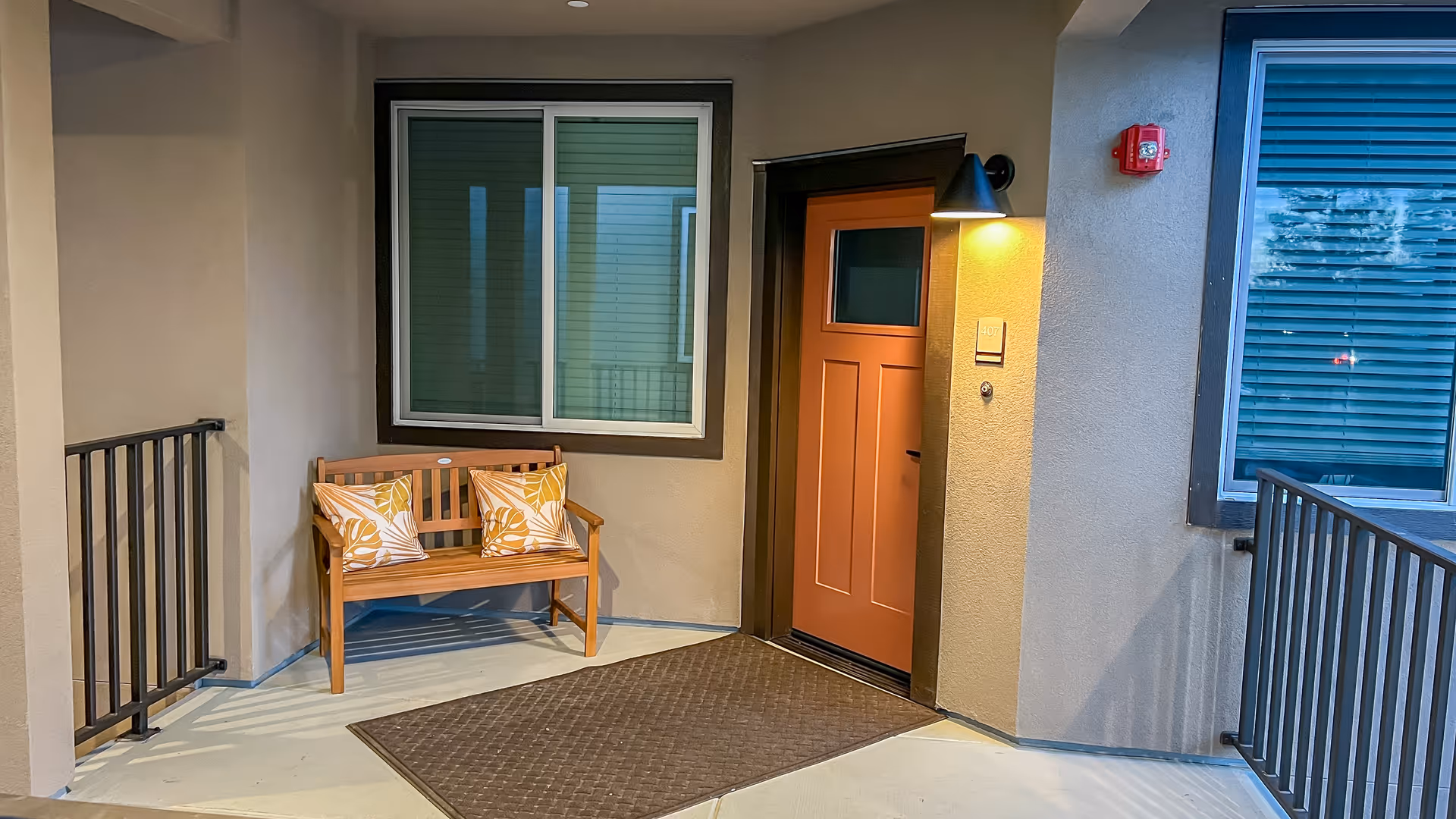 A covered exterior hallway with a wooden bench, orange door, window, and railing.