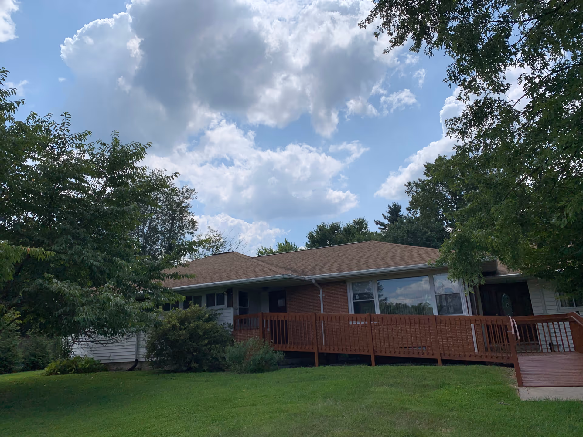 Single-story building with a brown roof and a wooden ramp leading to the entrance, surrounded by green grass and trees under a partly cloudy sky.