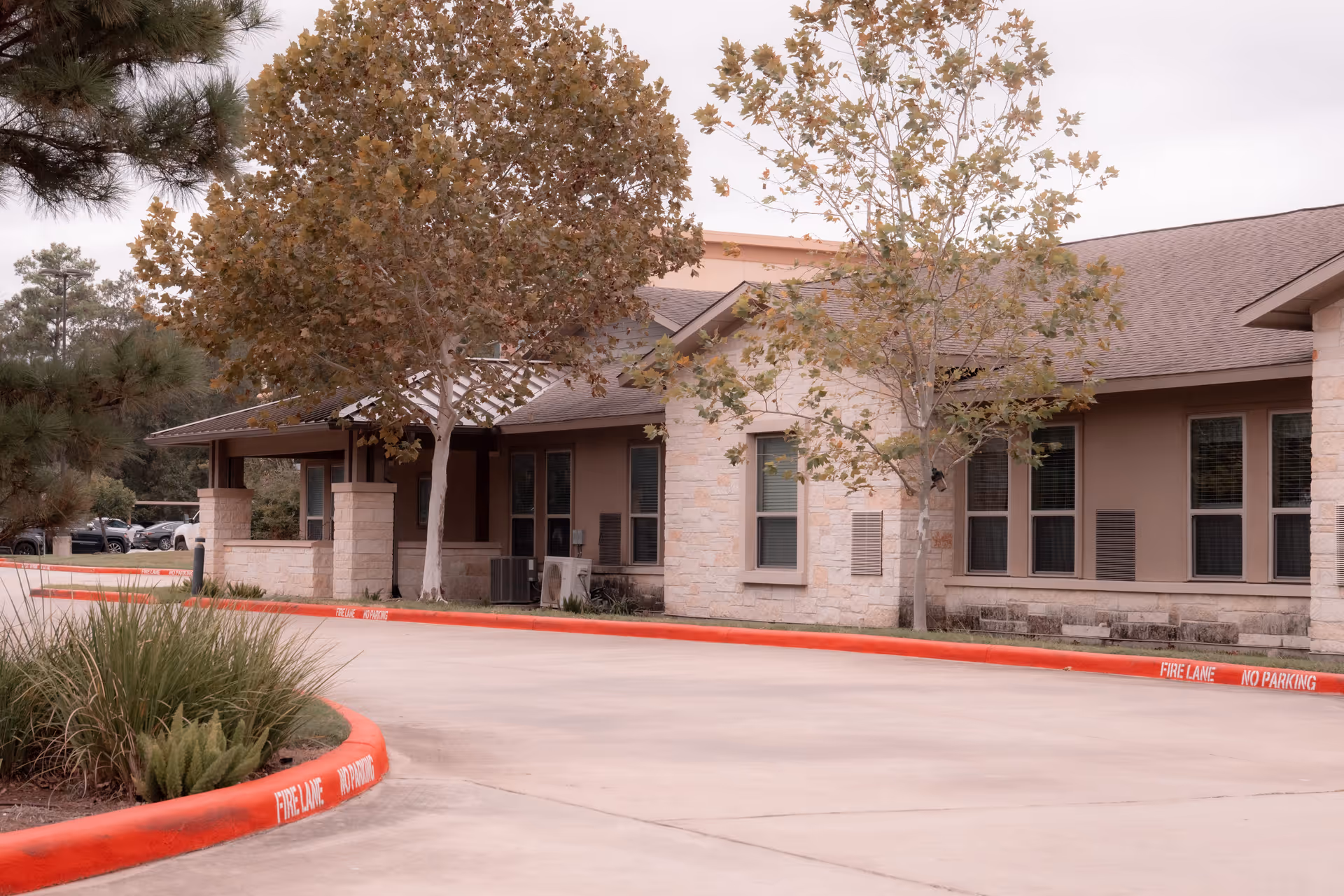 Exterior view of a single-story building with beige stone and brown walls, surrounded by trees with autumn leaves. A curved driveway with red-painted curbs marked 'FIRE LANE NO PARKING' is in the foreground, and several parked cars are visible in the background.