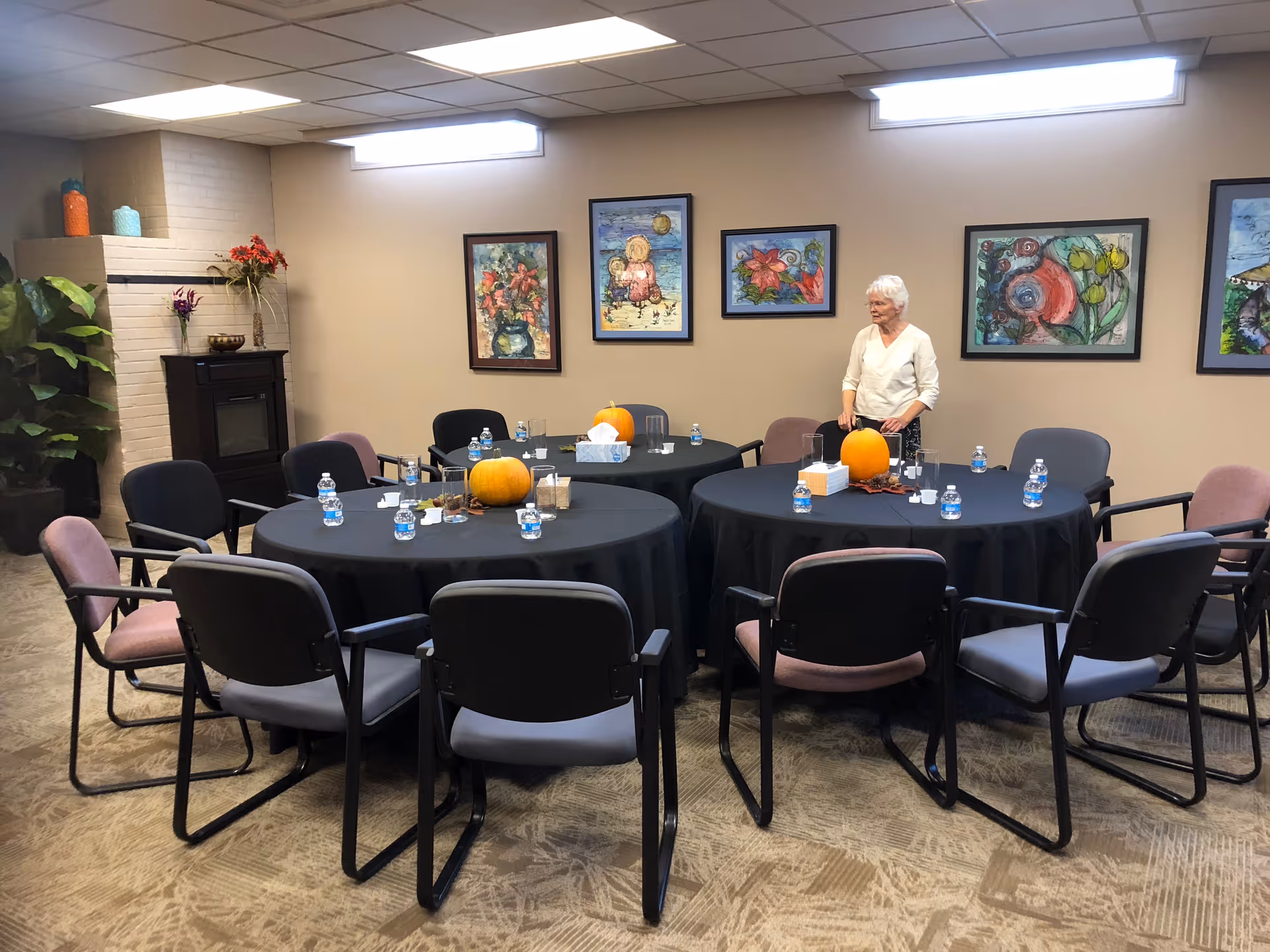 Senior woman standing by round dining tables with pumpkins in a decorated community dining room.