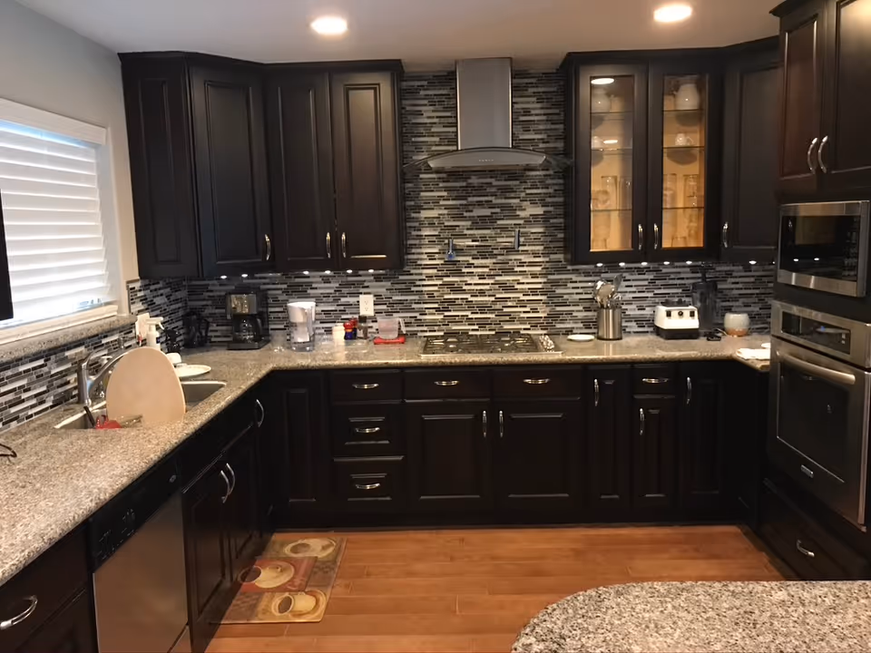 Modern kitchen with dark wood cabinets, granite countertops, and a mosaic tile backsplash. The kitchen features a stainless steel oven, microwave, dishwasher, and a range hood above the stove. There is a window with blinds on the left side, and various kitchen appliances and utensils are placed on the countertops.