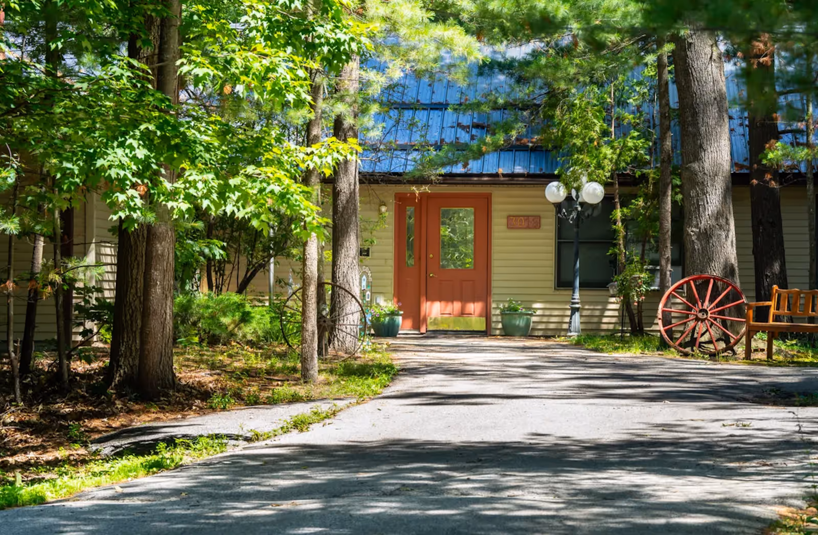 Tree-lined path to a beige building with a red door and a wooden wagon wheel.