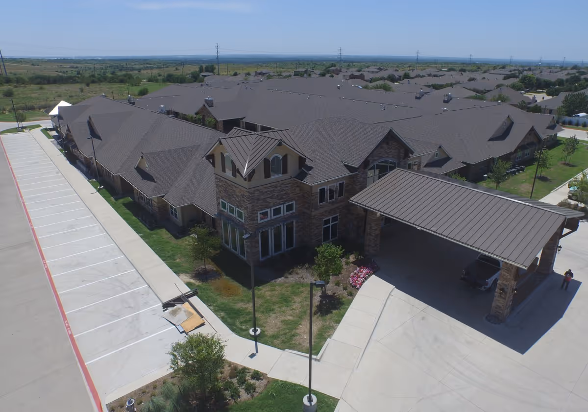 Aerial view of Bristol Park at Eagle Mountain Assisted Living & Memory Care building with a large covered entrance, multiple roof sections, surrounding greenery, and an adjacent empty parking lot.