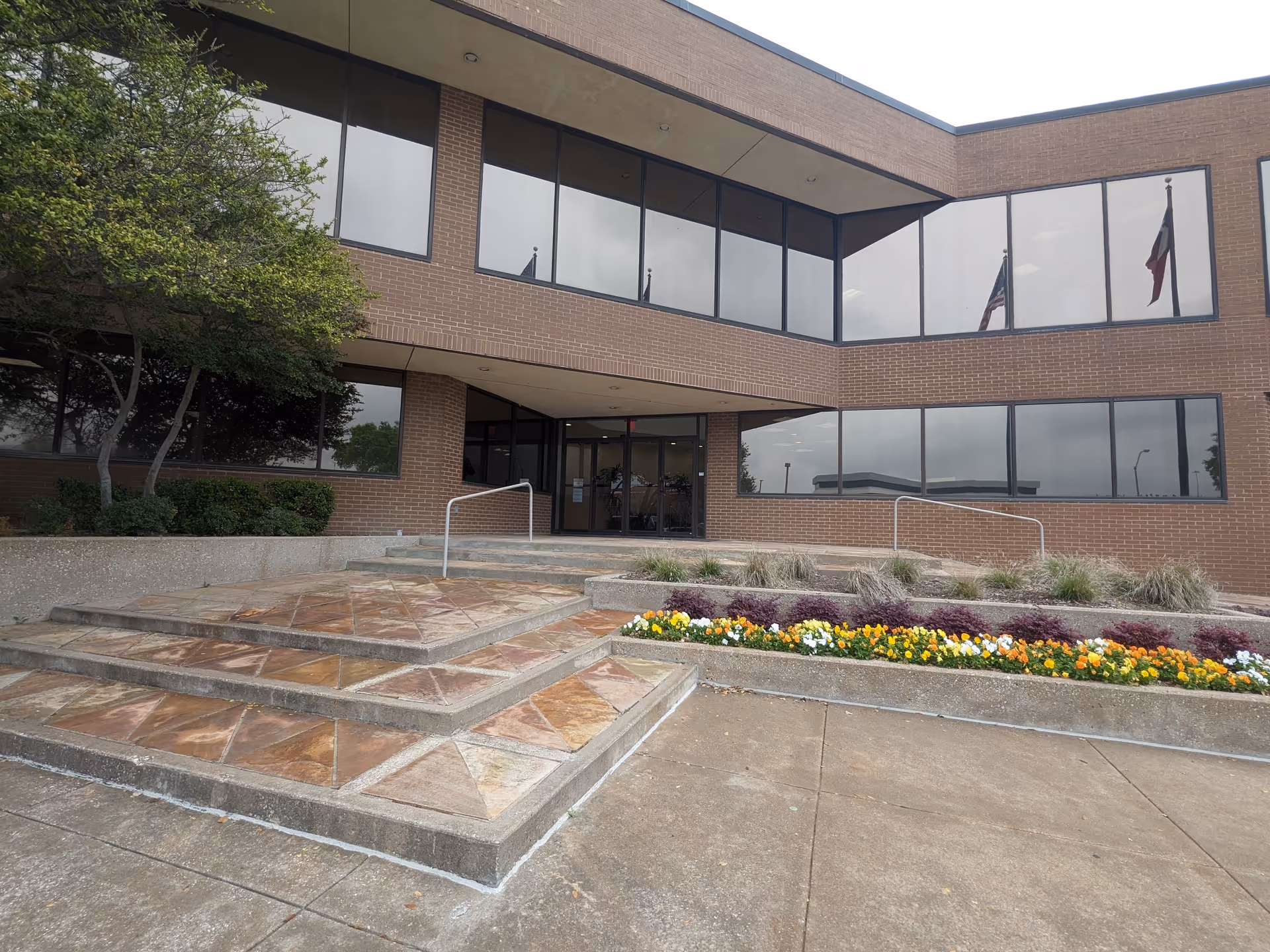 Brick senior care building with large windows, entrance stairs, and colorful flower beds.