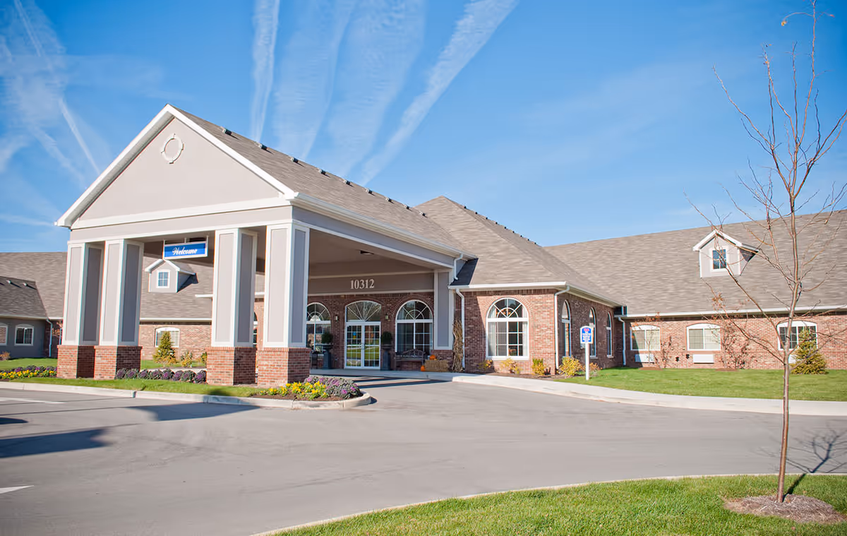 Brick senior living facility with porte-cochère entrance and manicured landscaping under blue sky.