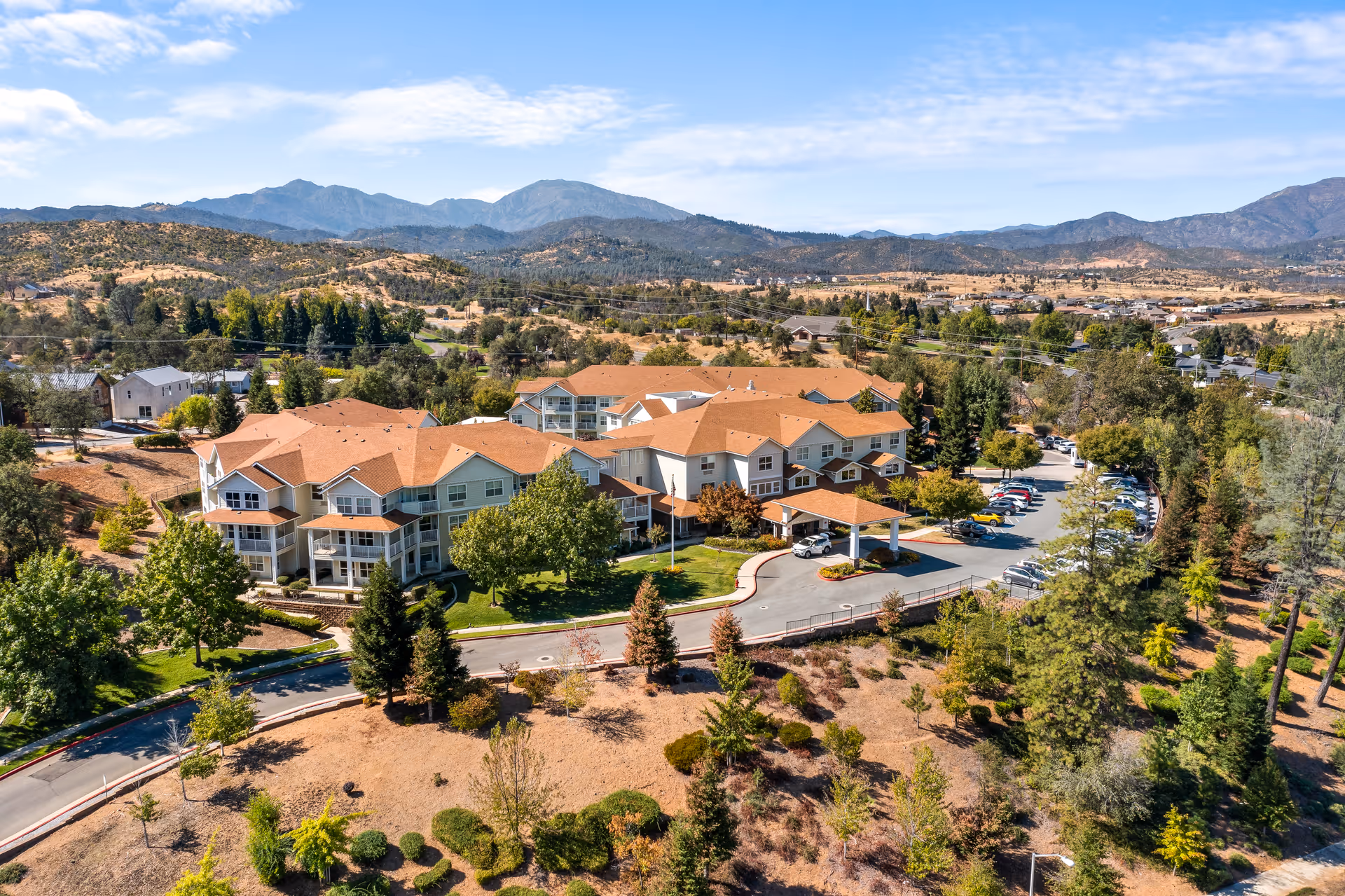 Aerial view of Shasta Estates Senior Living facility surrounded by trees and landscaping, with mountains in the background under a partly cloudy sky.