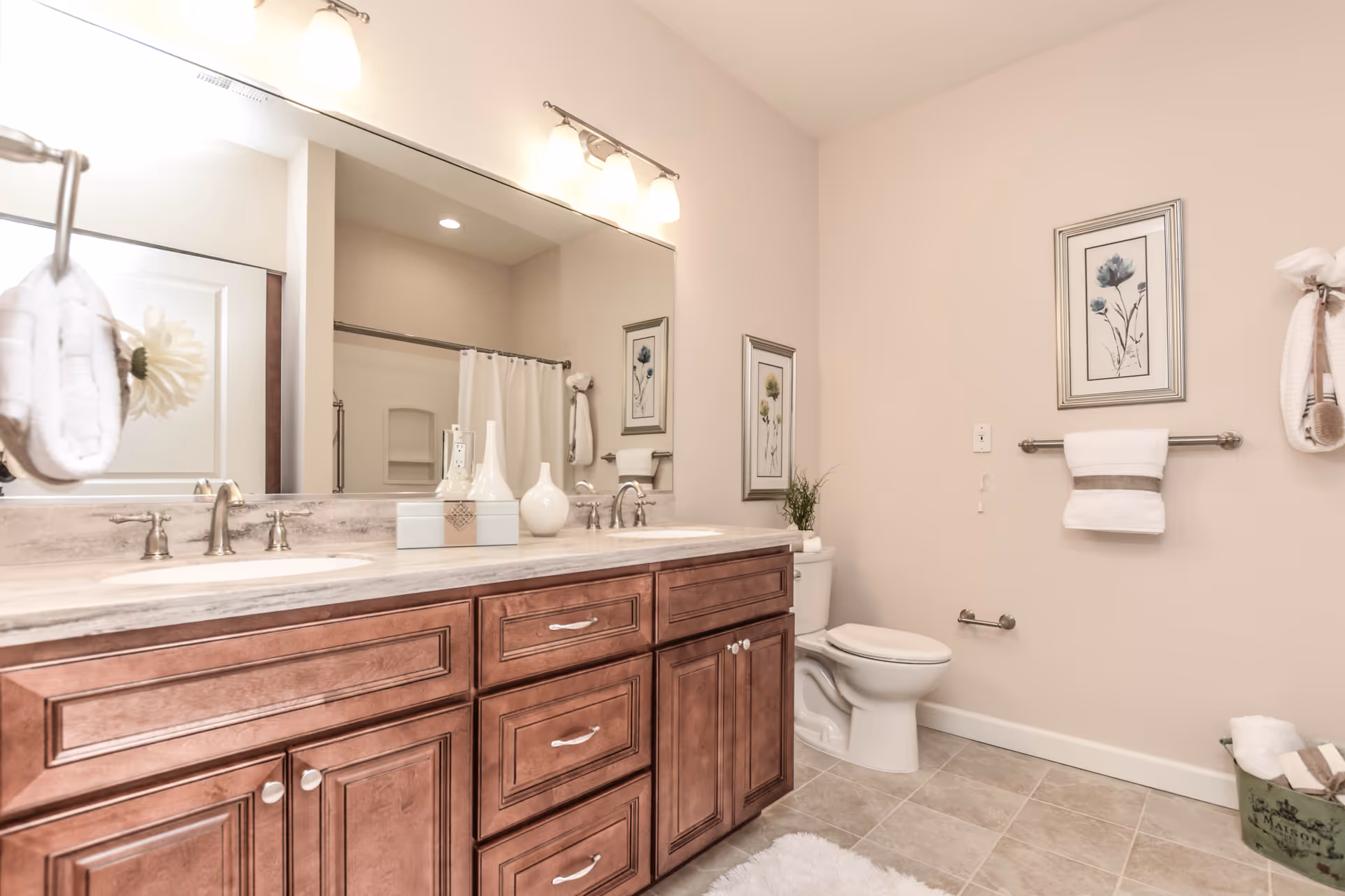 A clean and well-lit bathroom featuring a double sink vanity with wooden cabinets, a large mirror above the sinks, a toilet, and a shower with a white curtain. The walls are light-colored and decorated with framed floral artwork. Towels and decorative items are neatly arranged around the room.