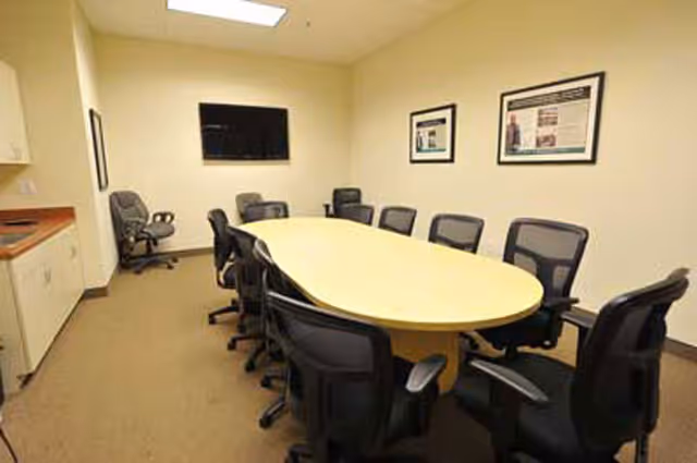 A conference room with a long oval table and multiple chairs, wall art, and a TV.