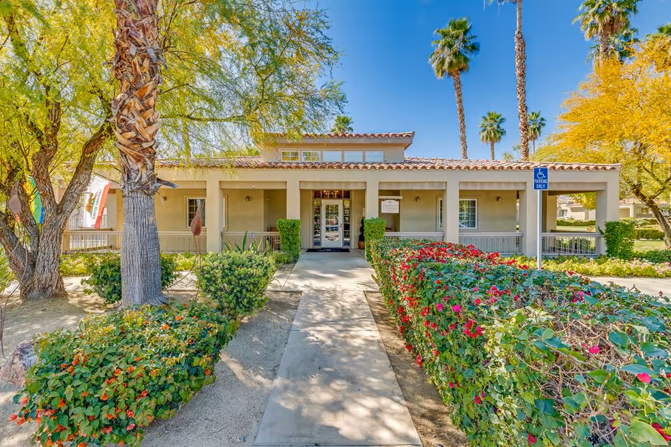 Front view of a single-story senior living facility with a walkway, shrubs, and palm trees.