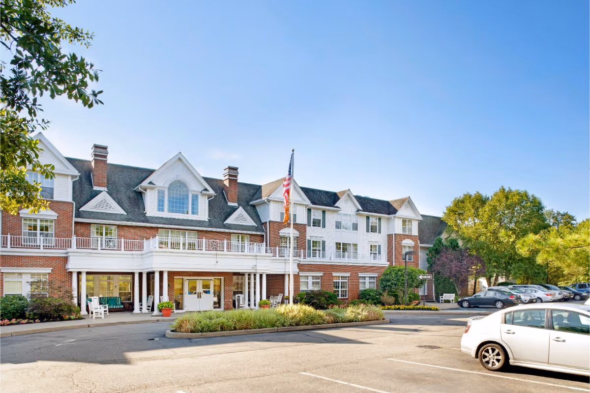 Front exterior of a multi-story brick and white senior living building with an entrance canopy, flagpole, landscaping, and parked cars.