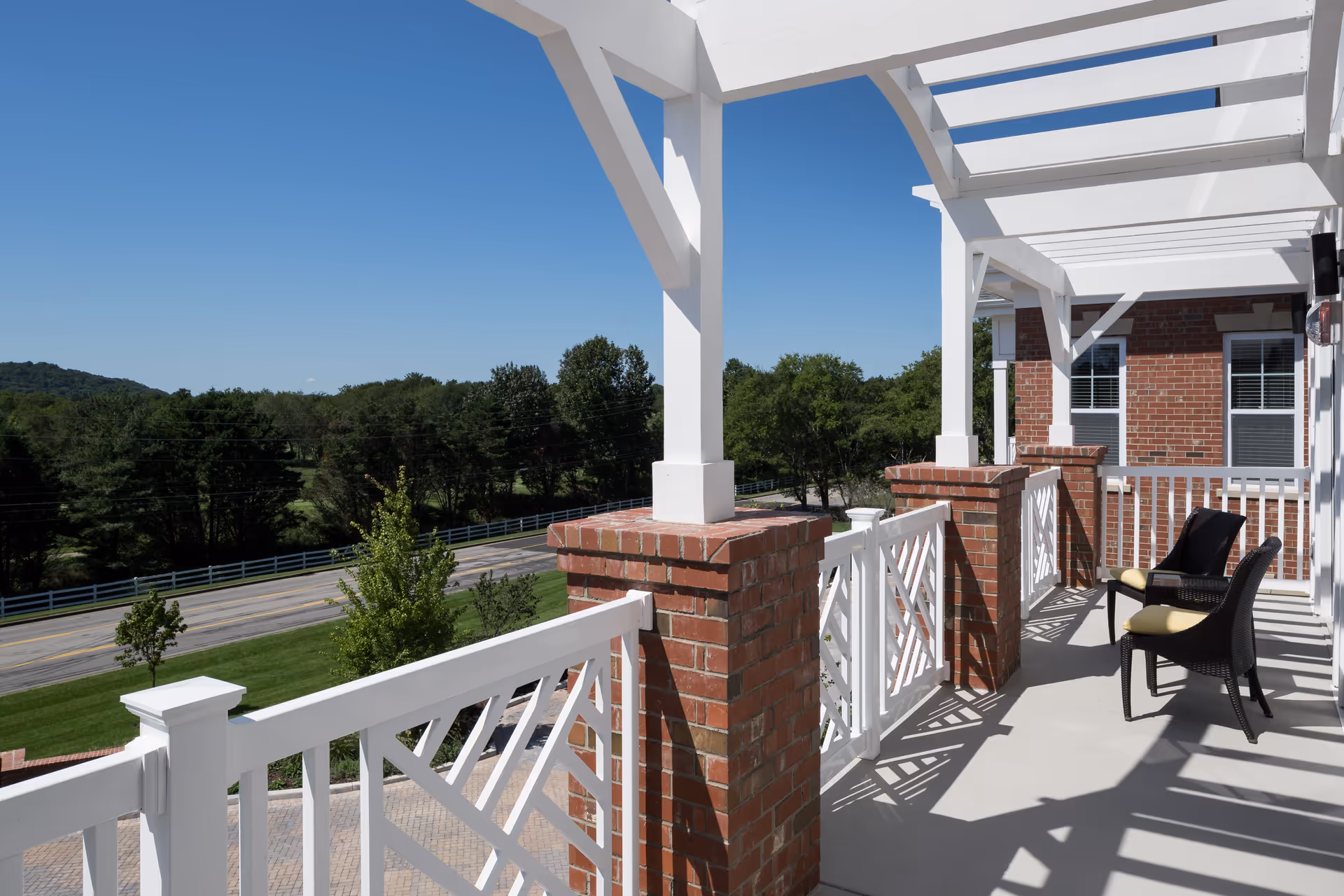 Sunlit brick-and-white balcony with chairs and a pergola-style roof overlooking a road and tree-lined landscape.