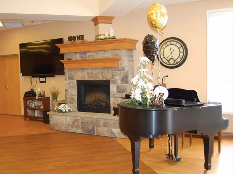A bright communal living area with a grand piano, stone fireplace, wall clock, and mounted television.