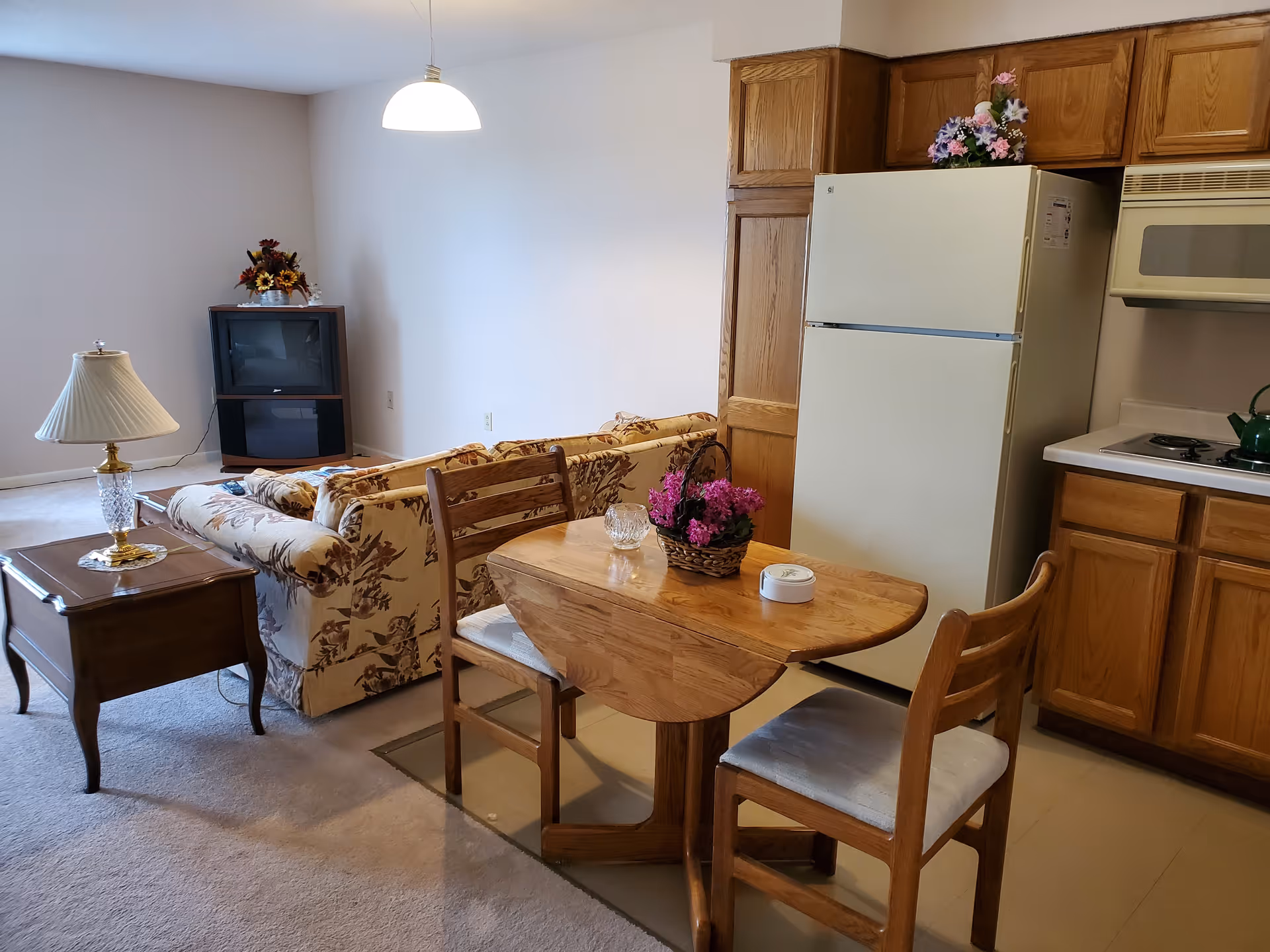 Open-plan living area with a floral sofa and TV, a small wooden dining table with chairs, and a kitchenette with refrigerator and cabinets.