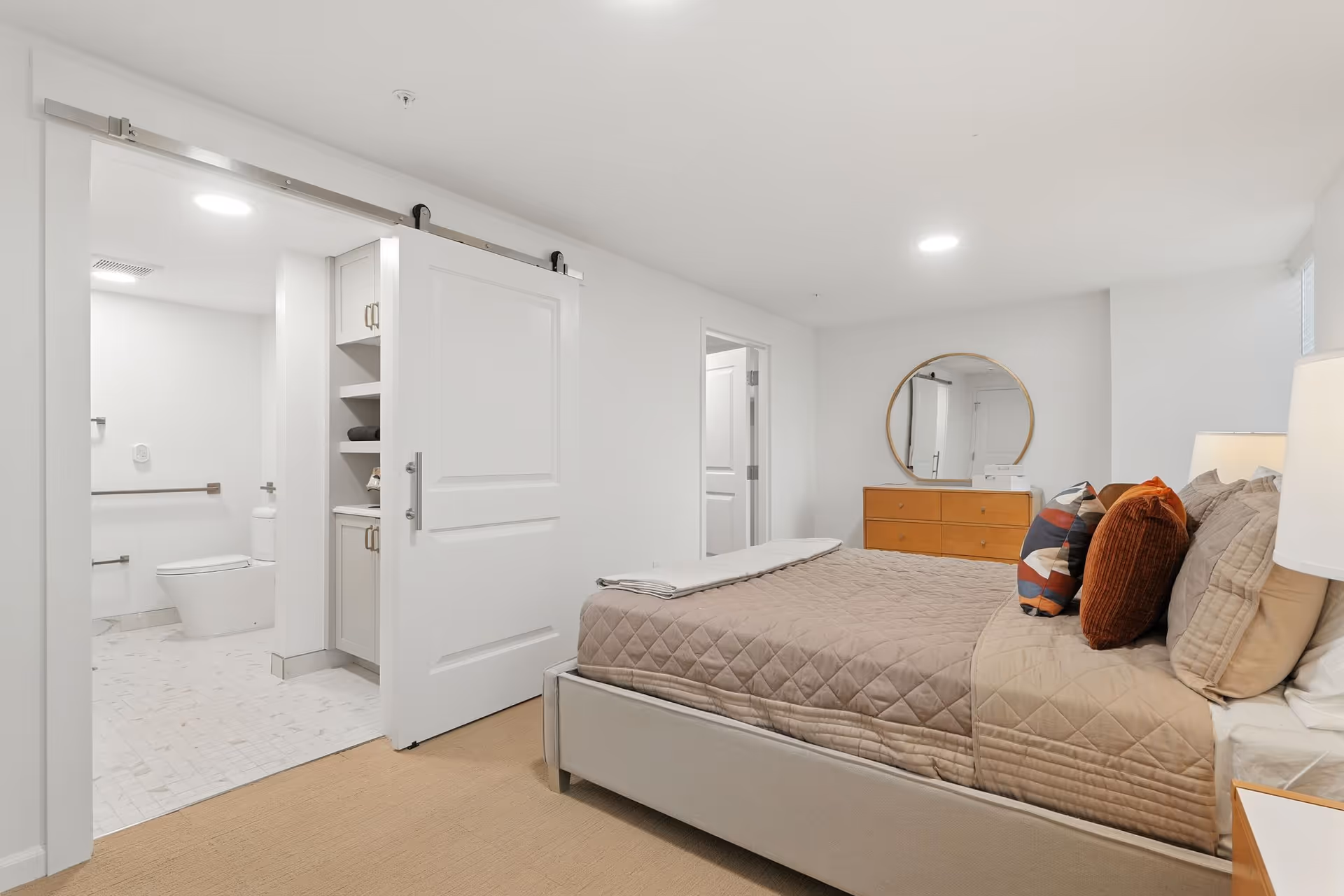 Bright bedroom with a bed, dresser, round mirror, and an open doorway to a tiled bathroom.