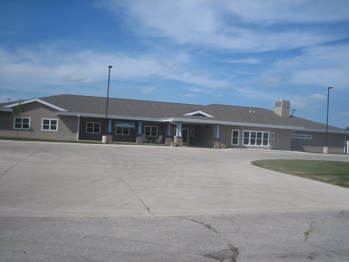 Single-story assisted living facility building with a large paved driveway under a blue sky.