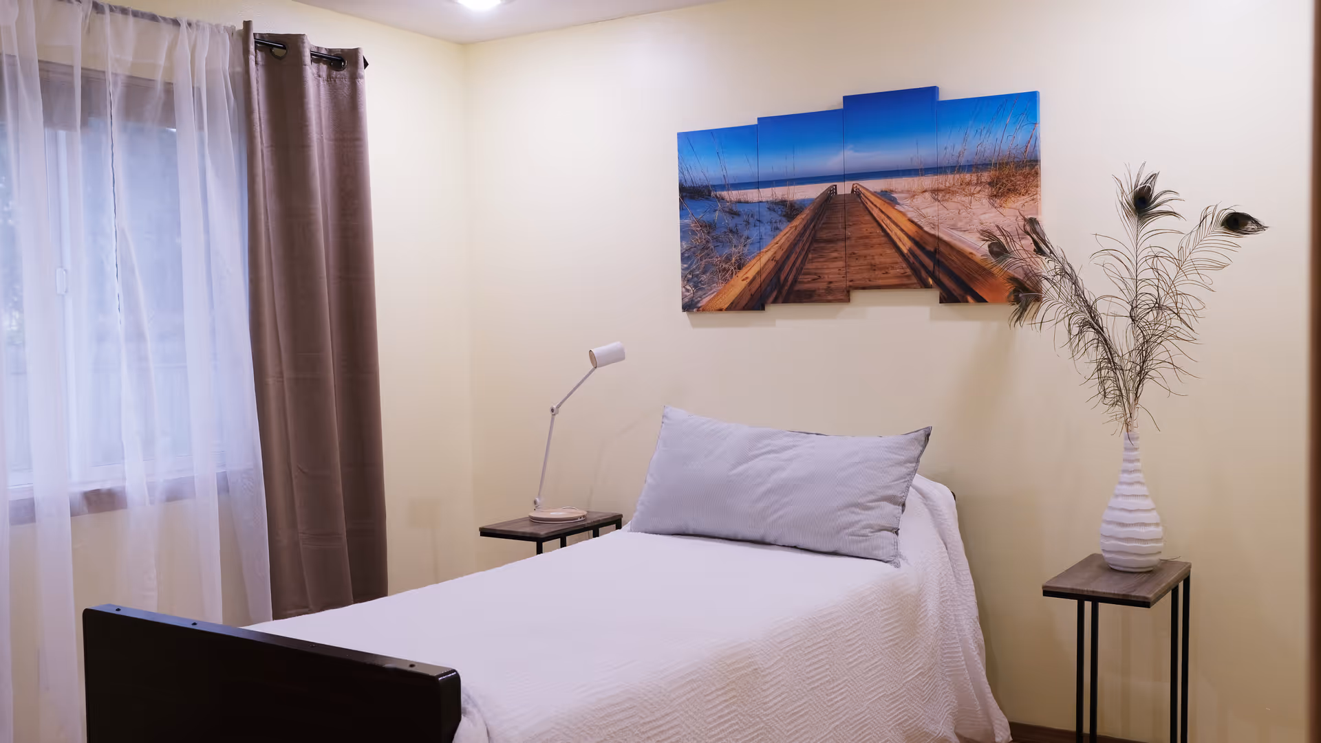 A tidy bedroom features a single bed, lamp, nightstand, window with curtains, and beach-themed wall art.